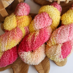 May include: Close-up of colorful yarn skeins in shades of pink, yellow, and peach. The yarn is arranged on a wooden surface, with a basket in the background. The yarn has a textured, looped appearance, ideal for knitting or crochet projects.