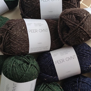 Puede incluir: Una colección de ovillos de lana Peer Gynt Tweed en varios colores, incluyendo marrón, verde y azul marino. Cada ovillo está etiquetado con el nombre de la marca y la palabra "Tweed". La lana está bien enrollada, mostrando una superficie texturizada.