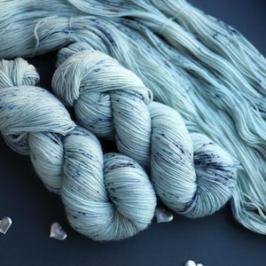 Può includere: Primo piano di diversi gomitoli di filato azzurro con macchie blu scuro. Il filato è disposto in modo sciolto e fluido, con alcuni gomitoli annodati. Ornamenti argentati a forma di cuore sono sparsi intorno al filato. Ideale per progetti di maglia e uncinetto.