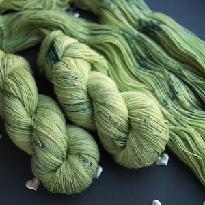 Può includere: Primo piano di matasse di filato verde e fili sciolti. Il filato è un verde chiaro, macchiettato, con accenti verde scuro. Il filato è disposto su una superficie scura, con piccole decorazioni argentate a forma di cuore.