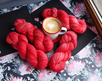 May include: A still-life image featuring vibrant red yarn skeins arranged around a white mug of coffee with a heart-shaped design in the foam. The scene is set on a floral patterned surface with decorative hearts.