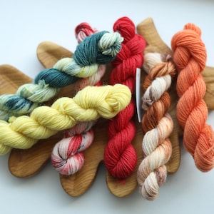 May include: A collection of six small yarn skeins in various colors including teal, yellow, red, orange, and variegated white and brown, arranged on a wooden board shaped like a large oak leaf with visible wood grain and smooth edges.