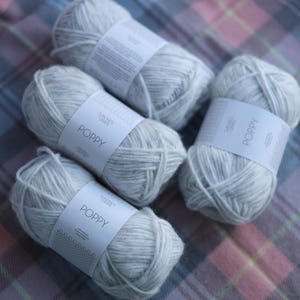 May include: Four skeins of light gray yarn labeled "POPPY" are arranged on a plaid fabric. The yarn is a soft, neutral color, ideal for knitting or crocheting projects. The labels are white with black text.