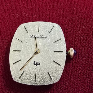 May include: A silver-toned Lucien Piccard watch face with black hour markers and hands. The watch has a square shape with rounded edges and a blue crown. The text "Lucien Piccard" and "LP" are visible on the face.