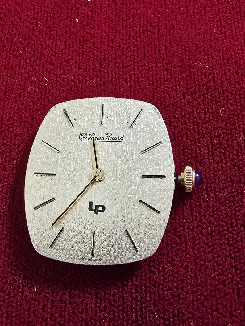May include: A silver-toned Lucien Piccard watch face with black hour markers and hands. The watch has a square shape with rounded edges and a blue crown. The text "Lucien Piccard" and "LP" are visible on the face.