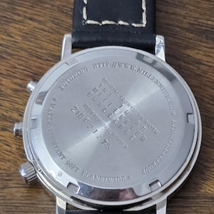 Op de afbeelding: Een zilverkleurige roestvrijstalen horlogekast met een zwarte leren band met witte stiksels. De achterkant is gegraveerd met tekst, waaronder "OFFICIAL COUNTDOWN WATCH", "THE THIRD MILLENNIUM CHALLENGE" en "WATER RESISTANT 90FT" en een nummer.