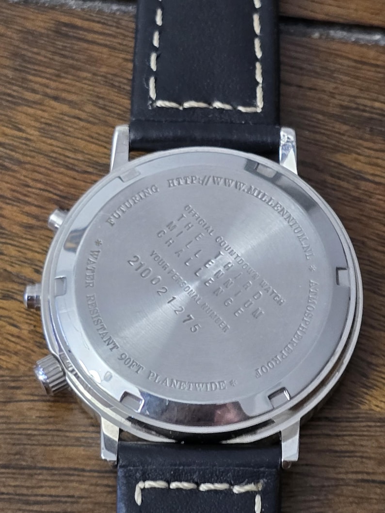 Op de afbeelding: Een zilverkleurige roestvrijstalen horlogekast met een zwarte leren band met witte stiksels. De achterkant is gegraveerd met tekst, waaronder "OFFICIAL COUNTDOWN WATCH", "THE THIRD MILLENNIUM CHALLENGE" en "WATER RESISTANT 90FT" en een nummer.