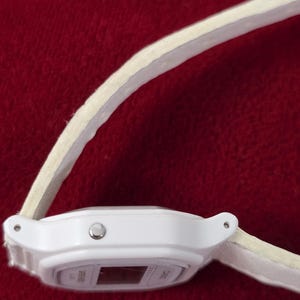 May include: A white digital watch with a light-colored band. The watch face is rectangular with a small display. The band appears to be made of a soft material. The watch is set against a red background.
