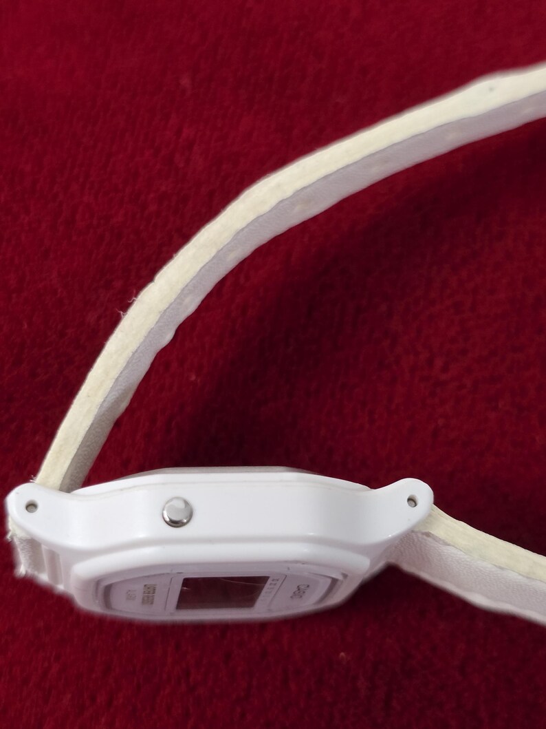 May include: A white digital watch with a light-colored band. The watch face is rectangular with a small display. The band appears to be made of a soft material. The watch is set against a red background.