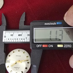 Puede incluir: Una esfera de reloj con manecillas y marcadores dorados se mide con un calibrador digital. El calibrador muestra 2,87 cm. La esfera del reloj tiene las palabras "electronic" y "Tradition" impresas.
