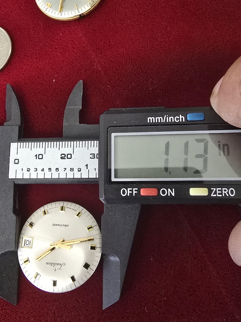 Puede incluir: Una esfera de reloj con manecillas y marcadores dorados se mide con un calibrador digital. El calibrador muestra 2,87 cm. La esfera del reloj tiene las palabras "electronic" y "Tradition" impresas.
