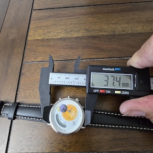 Op de afbeelding: Een polshorloge met een zwarte leren band met witte stiksels en een ronde witte wijzerplaat met een geel cirkelvormig midden, een paarse maanfase-indicator en een digitaal display. Een schuifmaat toont een kastdiameter van 37,4 mm.