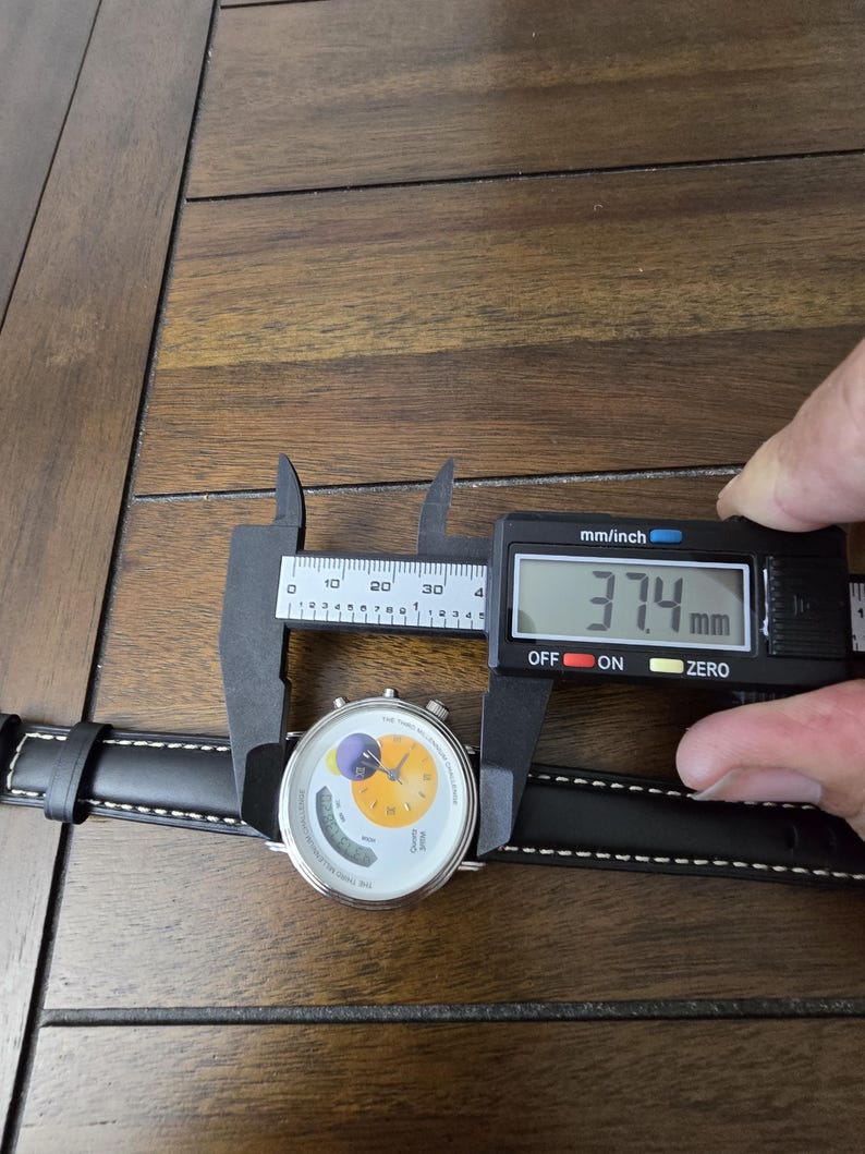Op de afbeelding: Een polshorloge met een zwarte leren band met witte stiksels en een ronde witte wijzerplaat met een geel cirkelvormig midden, een paarse maanfase-indicator en een digitaal display. Een schuifmaat toont een kastdiameter van 37,4 mm.