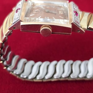 May include: A vintage Bulova wristwatch with a rectangular face and a gold-tone band. The watch features a light brown dial, gold hands, and a crown. The watch band is a combination of gold and silver-tone links, with red accents on the sides of the watch face.