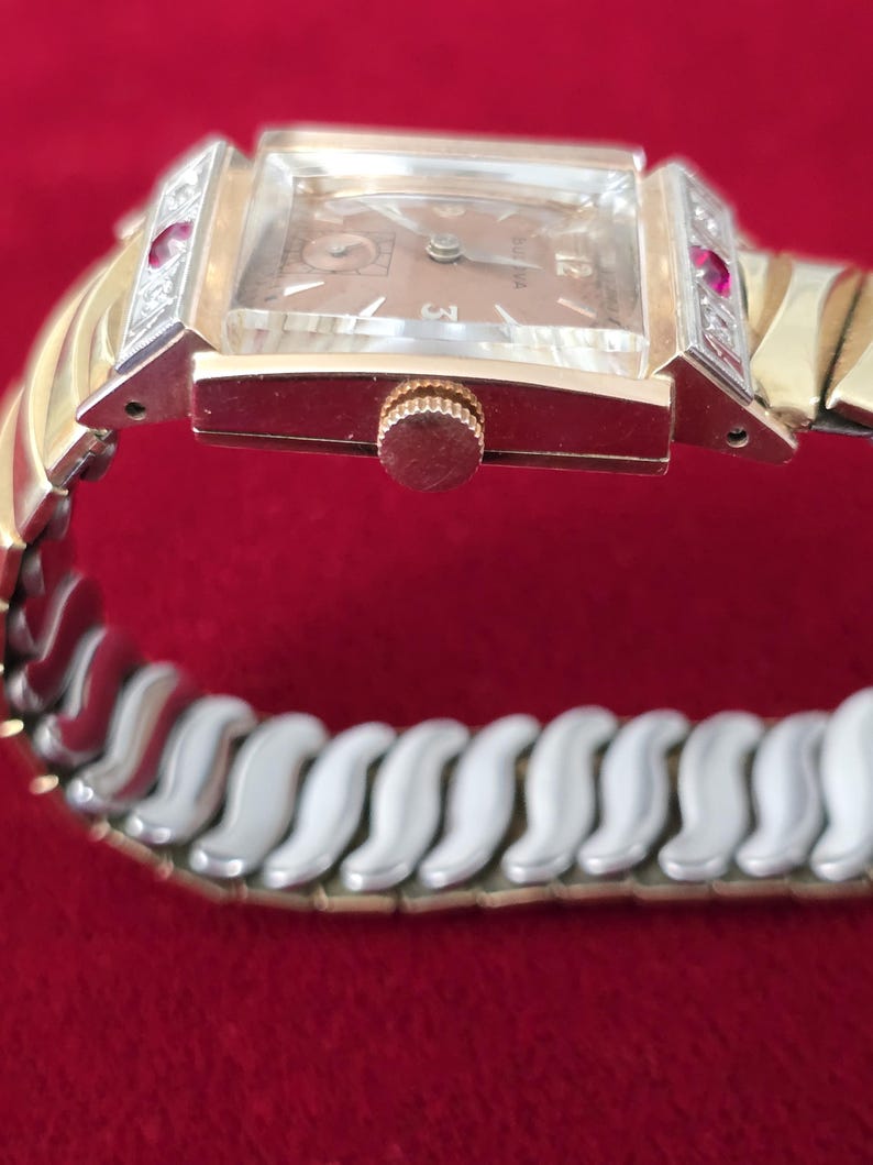 May include: A vintage Bulova wristwatch with a rectangular face and a gold-tone band. The watch features a light brown dial, gold hands, and a crown. The watch band is a combination of gold and silver-tone links, with red accents on the sides of the watch face.