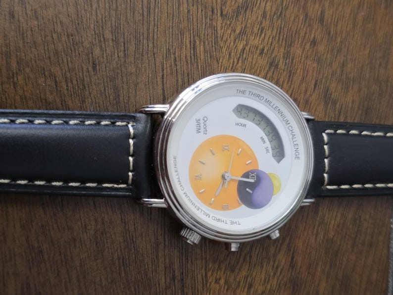 Op de afbeelding: Een zilverkleurig polshorloge met een zwarte leren band met witte stiksels. De witte wijzerplaat toont de tekst THE THIRD MILLENNIUM CHALLENGE, een digitaal display en een cirkelvormige oranje en paarse afbeelding met Romeinse cijfers.