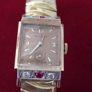 May include: A vintage Bulova wristwatch with a rectangular gold-tone case and a champagne-colored dial. The watch features diamond and ruby accents on the bezel and a gold-tone expansion band. The watch face displays the number 12, the Bulova brand name, and the numbers 3 and 9.