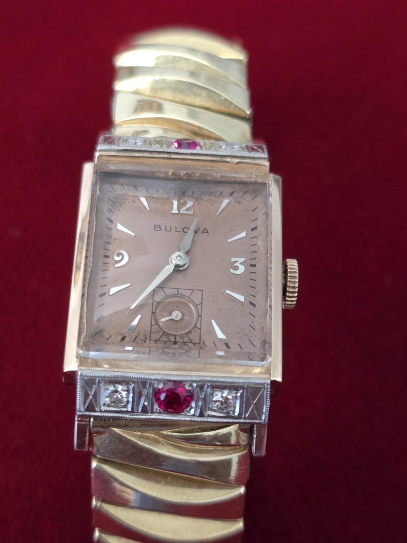 May include: A vintage Bulova wristwatch with a rectangular gold-tone case and a champagne-colored dial. The watch features diamond and ruby accents on the bezel and a gold-tone expansion band. The watch face displays the number 12, the Bulova brand name, and the numbers 3 and 9.