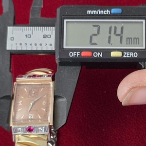 May include: A vintage Bulova watch with a rectangular face, gold-tone band, and a light brown dial. The watch is being measured with a digital caliper, showing a width of 21.4 mm. The watch features red and clear gemstones.