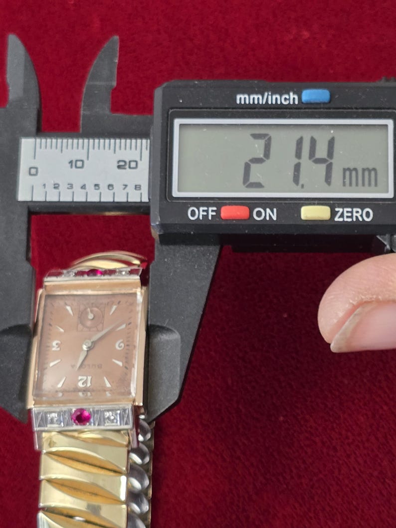 May include: A vintage Bulova watch with a rectangular face, gold-tone band, and a light brown dial. The watch is being measured with a digital caliper, showing a width of 21.4 mm. The watch features red and clear gemstones.