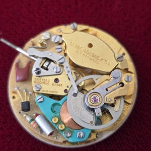 Puede incluir: Primer plano de un mecanismo de reloj con engranajes dorados, un componente turquesa y el texto "THIRTEEN 13 JEWELS SWISS". El mecanismo del reloj se encuentra sobre un fondo burdeos.