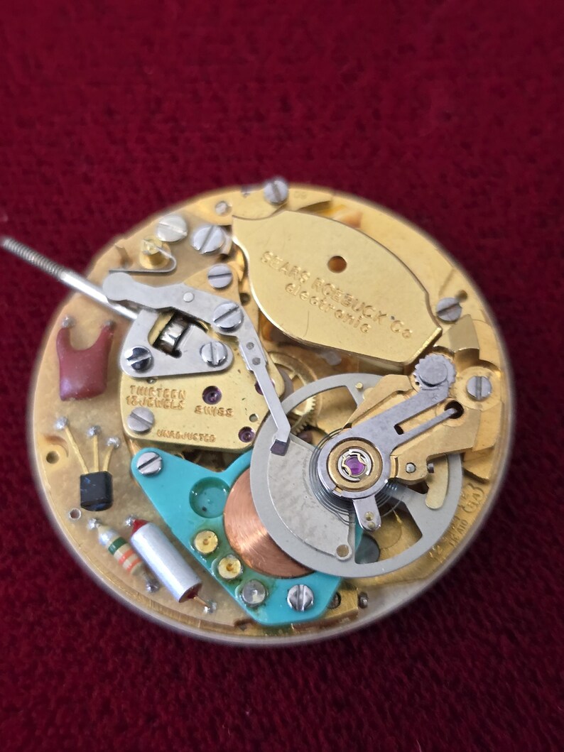 Puede incluir: Primer plano de un mecanismo de reloj con engranajes dorados, un componente turquesa y el texto "THIRTEEN 13 JEWELS SWISS". El mecanismo del reloj se encuentra sobre un fondo burdeos.