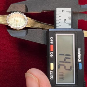 K&ouml;nnte beinhalten: Eine goldfarbene Armbanduhr mit diamantbesetzter L&uuml;nette und einem goldenen Mesh-Armband wird mit einem digitalen Messschieber gemessen. Die Digitalanzeige zeigt 14,2 mm an. Das Zifferblatt ist oval und hat goldene Zeiger.