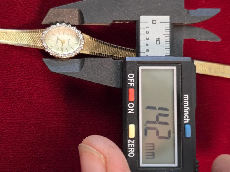 K&ouml;nnte beinhalten: Eine goldfarbene Armbanduhr mit diamantbesetzter L&uuml;nette und einem goldenen Mesh-Armband wird mit einem digitalen Messschieber gemessen. Die Digitalanzeige zeigt 14,2 mm an. Das Zifferblatt ist oval und hat goldene Zeiger.
