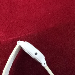 May include: A white wristwatch with a white band. The watch face is rectangular with a silver-colored button. The band is made of a soft, textured material. The watch is set against a red background.