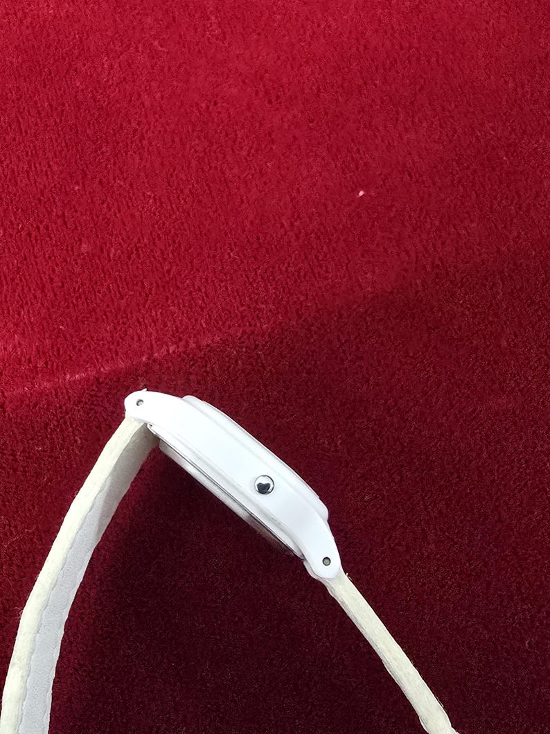 May include: A white wristwatch with a white band. The watch face is rectangular with a silver-colored button. The band is made of a soft, textured material. The watch is set against a red background.