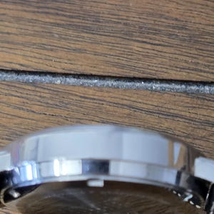 Op de afbeelding: Een zijaanzicht van een ronde, zilverkleurige horlogekast met een gepolijste metalen afwerking. De ronde behuizing is voorzien van een kleine witte kroon die aan de zijkant uitsteekt en de achterkant van het horloge is zichtbaar met een donker oppervlak.
