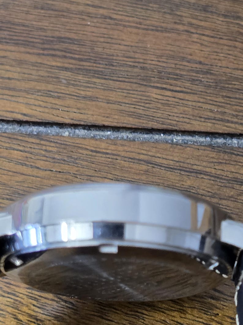 Op de afbeelding: Een zijaanzicht van een ronde, zilverkleurige horlogekast met een gepolijste metalen afwerking. De ronde behuizing is voorzien van een kleine witte kroon die aan de zijkant uitsteekt en de achterkant van het horloge is zichtbaar met een donker oppervlak.