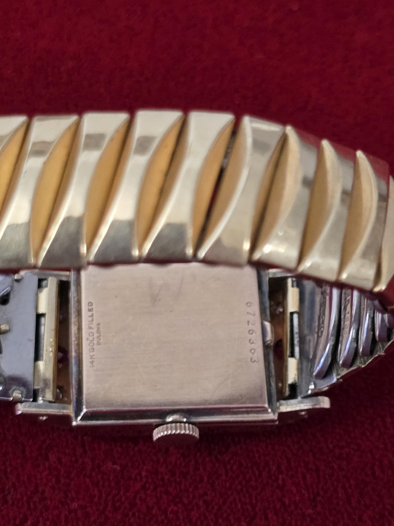 May include: Close-up of a vintage gold-filled Bulova watch. The watch features a rectangular face and a gold-toned, expandable band. The back of the watch is visible, with the inscription "14K Gold Filled Bulova" and the number "6726303".