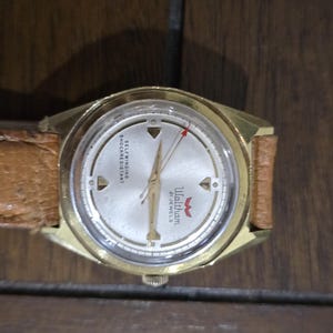 May include: A vintage Waltham wristwatch with a gold-tone case and a light-colored face. The watch features a brown leather band and a red second hand. The watch face has the text "Waltham 21 Jewels".