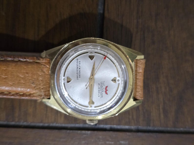 May include: A vintage Waltham wristwatch with a gold-tone case and a light-colored face. The watch features a brown leather band and a red second hand. The watch face has the text "Waltham 21 Jewels".