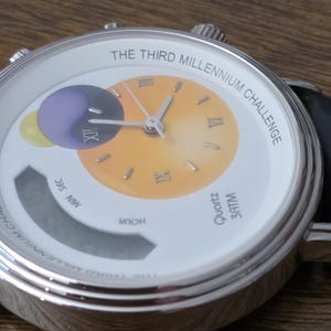 Op de afbeelding: Een zilverkleurig polshorloge met een witte wijzerplaat met de tekst THE THIRD MILLENNIUM CHALLENGE. De wijzerplaat heeft een geel verloop in het midden, Romeinse cijfers, een paars en geel cirkelvormig grafisch element en een digitaal display.