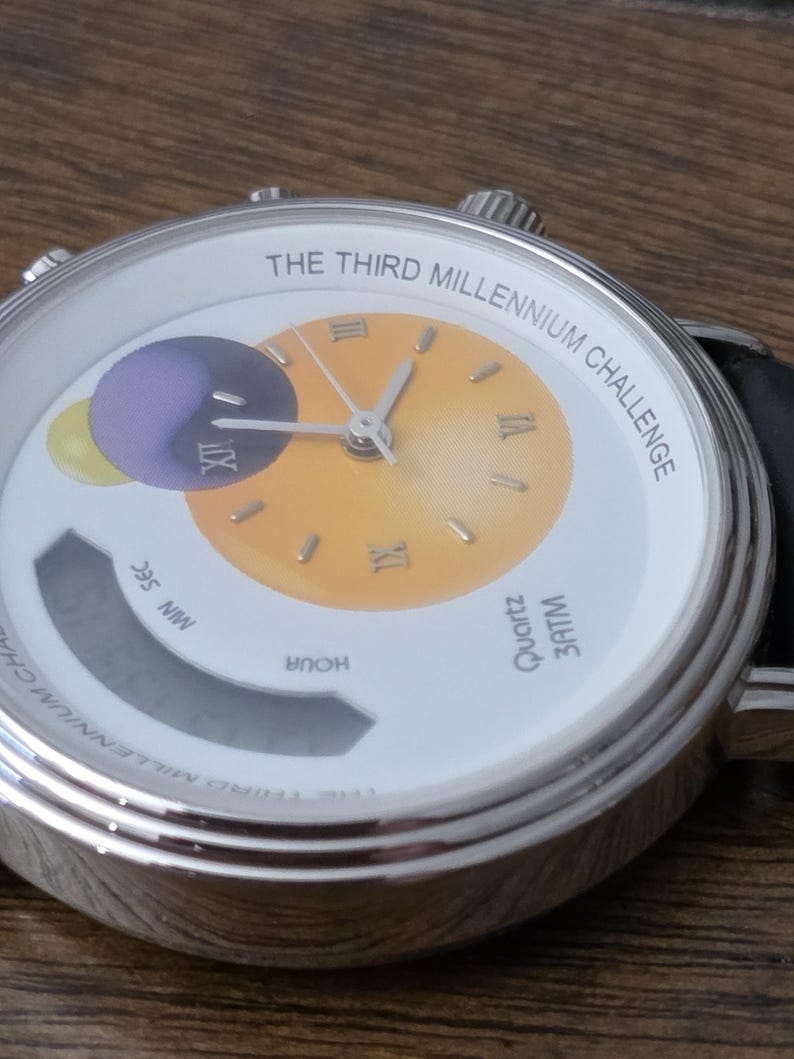 Op de afbeelding: Een zilverkleurig polshorloge met een witte wijzerplaat met de tekst THE THIRD MILLENNIUM CHALLENGE. De wijzerplaat heeft een geel verloop in het midden, Romeinse cijfers, een paars en geel cirkelvormig grafisch element en een digitaal display.