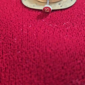 May include: Close-up of a watch movement with a gold-colored base and silver gears. The watch stem is visible, topped with a red Omega logo. The background is a textured red surface.