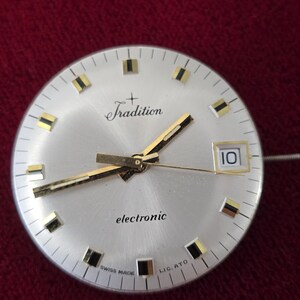 Puede incluir: Esfera de reloj plateada con manecillas e indicadores dorados. La palabra "Tradition" est&aacute; encima del centro, y "electronic" debajo. La fecha se muestra como "10". Tambi&eacute;n son visibles las palabras "SWISS MADE" y "LIG. ATO".