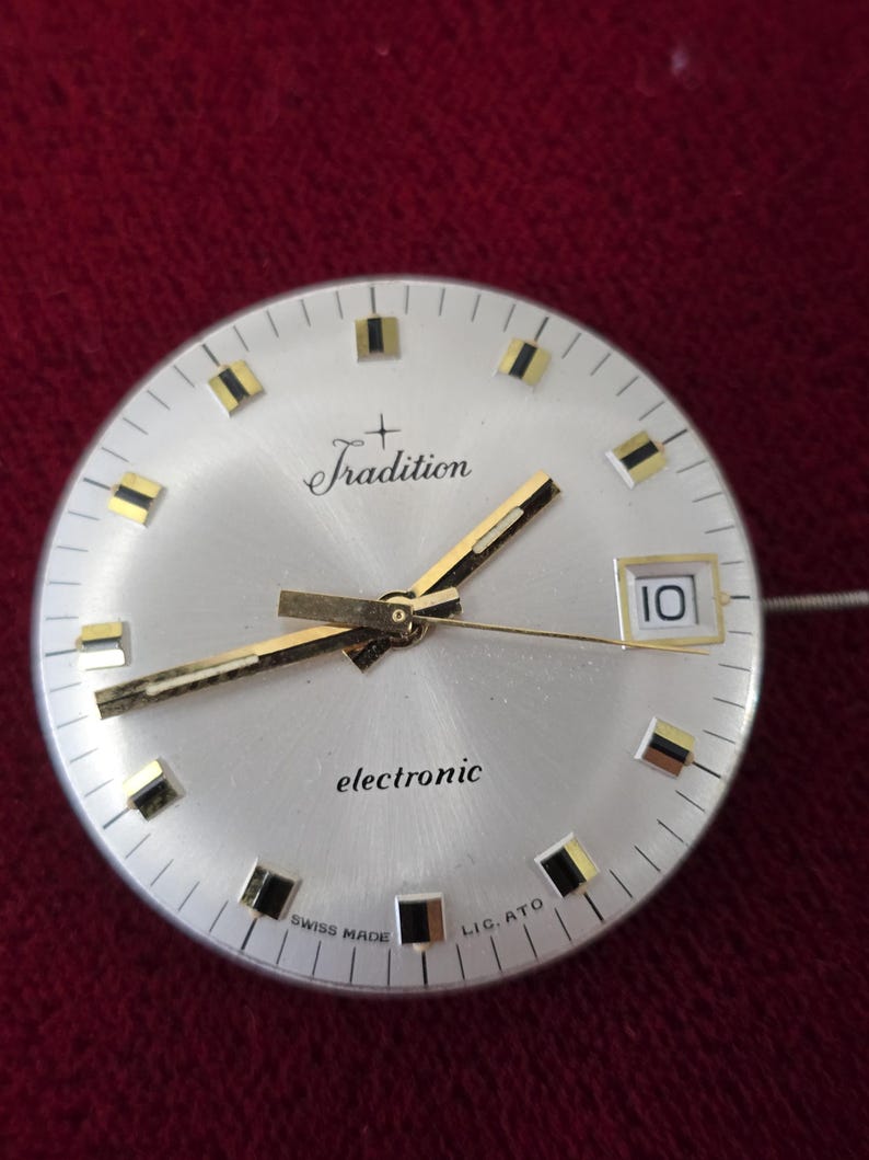 Puede incluir: Esfera de reloj plateada con manecillas e indicadores dorados. La palabra "Tradition" est&aacute; encima del centro, y "electronic" debajo. La fecha se muestra como "10". Tambi&eacute;n son visibles las palabras "SWISS MADE" y "LIG. ATO".