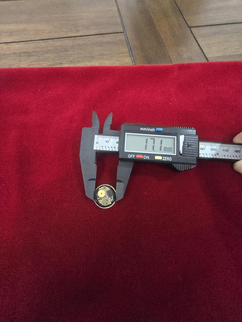 K&ouml;nnte beinhalten: Ein kleiner, runder, goldfarbener Knopf mit einem schwarz-goldenen Yin-Yang-Design, gemessen mit einem digitalen Messschieber von 1,71 cm. Der Knopf liegt auf rotem Samthintergrund.