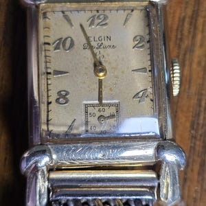 May include: A rectangular Elgin De Luxe wristwatch with a gold-tone case and a metallic mesh band. The off-white dial features gold-tone Arabic numerals at 12, 10, 8, 4, and 2, with a small sub-dial for seconds at the bottom and a textured bezel.