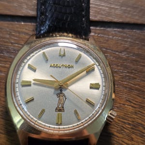 May include: A gold-tone Accutron wristwatch with a silver dial, gold-tone hour markers, and gold-tone hands. The dial features a small graphic of a standing figure near the six o'clock position and a black textured leather watch strap.