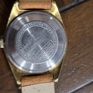 May include: Close-up of a vintage gold-toned watch with a brown leather strap. The watch face is not visible, but the back is detailed with text that reads "Antimagnetic Shock Resistant Waterproof" and the brand name "Waltham".