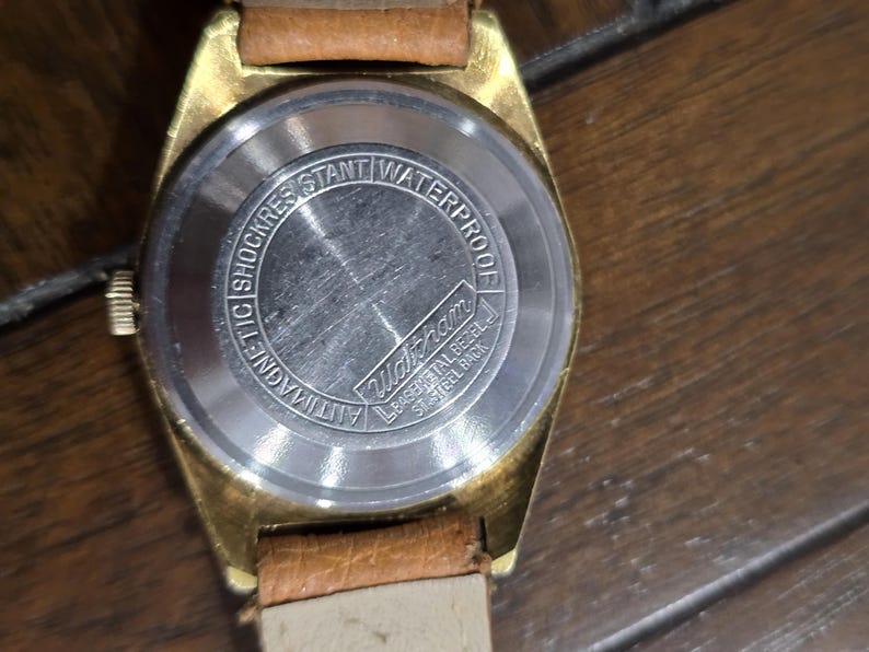 May include: Close-up of a vintage gold-toned watch with a brown leather strap. The watch face is not visible, but the back is detailed with text that reads "Antimagnetic Shock Resistant Waterproof" and the brand name "Waltham".