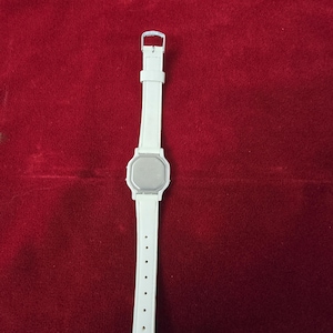 May include: A white digital watch with a rectangular face and a white strap. The watch has a silver buckle and a series of holes for adjustment. The watch is displayed against a red background.