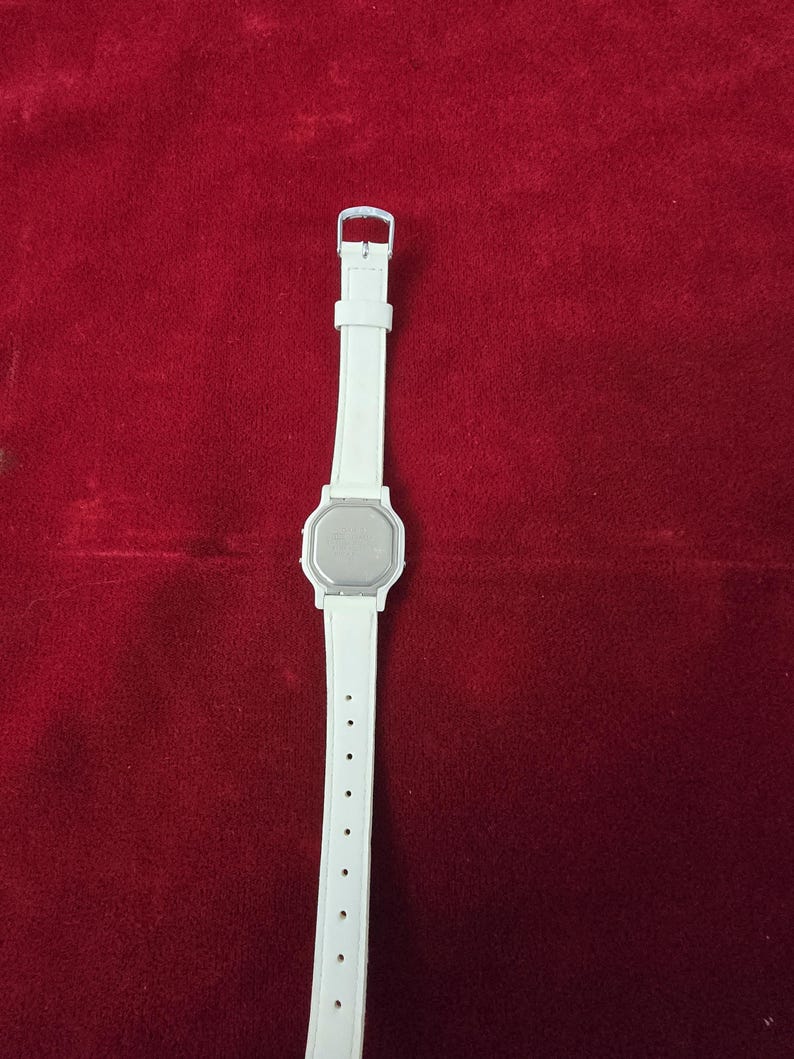 May include: A white digital watch with a rectangular face and a white strap. The watch has a silver buckle and a series of holes for adjustment. The watch is displayed against a red background.