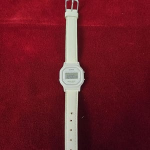 May include: A white Casio digital watch with a matching white leather strap. The watch face displays the time. The strap has a silver buckle and multiple adjustment holes. The watch is a classic, minimalist design.