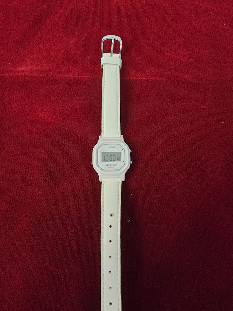 May include: A white Casio digital watch with a matching white leather strap. The watch face displays the time. The strap has a silver buckle and multiple adjustment holes. The watch is a classic, minimalist design.