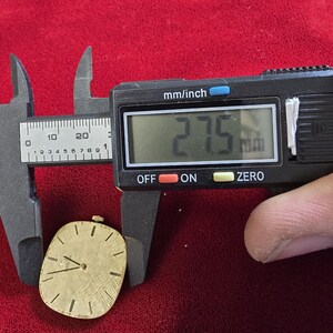 May include: A gold-toned watch face is measured with a digital caliper. The caliper displays 27.5 mm. The watch face has a textured surface, black hands, and black hour markers. The watch face is oval-shaped and sits on a red surface.